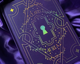 May include: A purple hardcover book featuring gold foil detailing on the front cover. The design includes a central keyhole shape, intricate geometric borders, vine motifs, and the text CLAVIS AUREA. Small star symbols are scattered throughout.