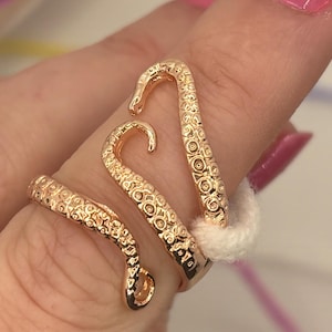 May include: A gold-colored ring wraps around a finger, featuring a design resembling octopus tentacles. The ring has detailed textures and curves, with a polished finish. The tentacles are arranged to create a unique and eye-catching jewelry piece.