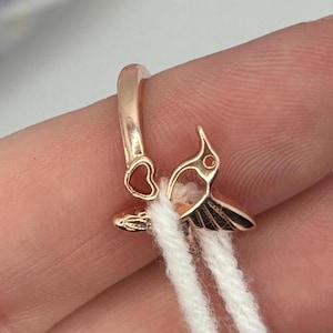 May include: A rose gold hummingbird charm with black wing details and a small red accent. The charm is attached to a white cord, and the design includes a heart-shaped element.