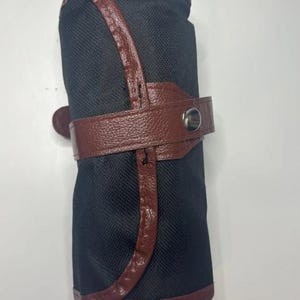Pode incluir: Um estojo de l&aacute;pis preto com uma al&ccedil;a de couro marrom e um fecho de press&atilde;o. O estojo &eacute; enrolado e tem uma guarni&ccedil;&atilde;o de couro marrom.