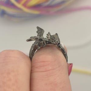 May include: A silver dragon ring with detailed wings and a textured body. The dragon is perched on the ring's band, creating a unique design. The ring is held up by a finger, showcasing the intricate craftsmanship. A whimsical piece of jewellery.