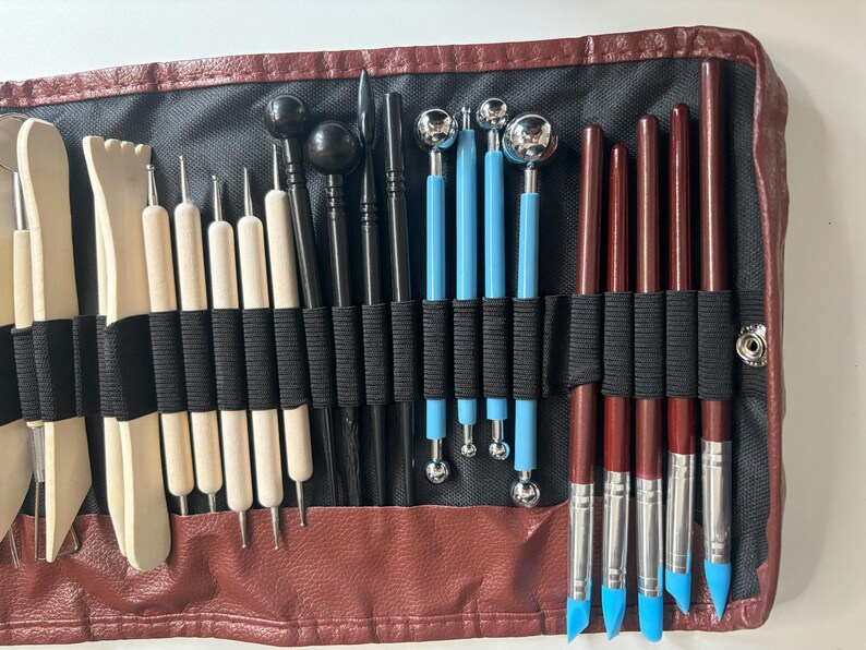 May include: A brown leather case with a black interior holds a variety of tools for sculpting and crafting. The tools include wooden, metal, and plastic implements with different shapes and sizes.