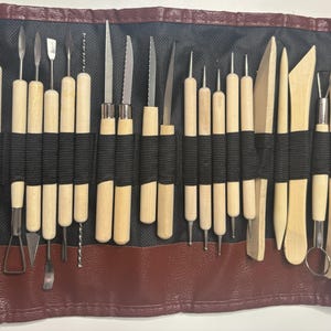 Pode incluir: Um conjunto de 25 ferramentas de cer&acirc;mica em um rolo de couro marrom. As ferramentas s&atilde;o feitas de madeira e metal e s&atilde;o usadas para moldar e esculpir a argila.