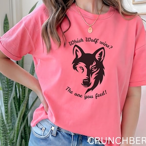 May include: A coral-coloured t-shirt with a black wolf graphic and the text "Which Wolf wins? The one you feed!" The shirt has short sleeves and a relaxed fit, suitable for casual wear.