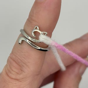 Puede incluir: Un anillo de plata con un diseño de gato. El anillo está en un dedo y tiene dos hebras de hilo, una blanca y una rosa, envueltas alrededor.