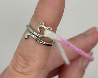 Könnte beinhalten: Ein silberner Ring mit einem Katzenmotiv. Der Ring wird an einem Finger getragen und hat zwei Fäden Garn, einen weißen und einen rosa, die um ihn herumgewickelt sind.