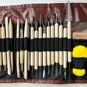 May include: A brown leather roll-up case with a black elastic band closure holds a set of 30 assorted sculpting tools. The tools have wooden handles and metal tips. The case also includes two yellow sponges and a small brush.