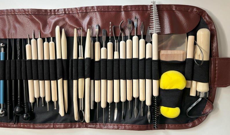 May include: A brown leather roll-up case with a black elastic band closure holds a set of 30 assorted sculpting tools. The tools have wooden handles and metal tips. The case also includes two yellow sponges and a small brush.
