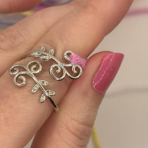 Puede incluir: Un anillo plateado con un diseño floral. El anillo presenta patrones delicados y arremolinados y pequeños detalles de hojas. El anillo se lleva en un dedo con esmalte de uñas rosa.