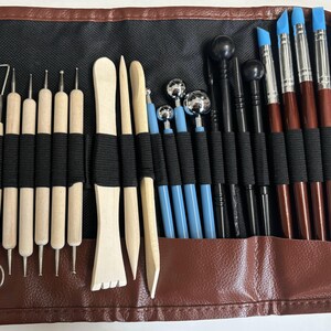 May include: A brown leather roll-up case with a black fabric interior holds a set of 28 sculpting tools. The tools include wooden sculpting tools, metal sculpting tools, and silicone sculpting tools. The tools are arranged in rows and are secured with black elastic bands.