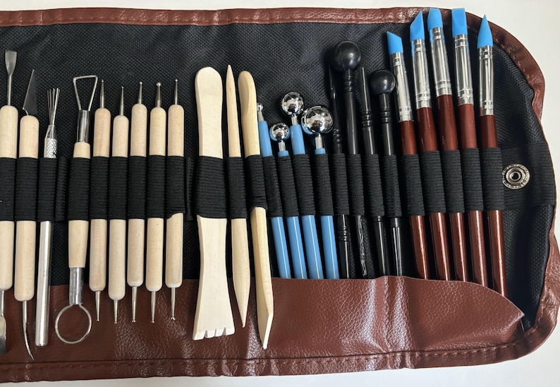 May include: A brown leather roll-up case with a black fabric interior holds a set of 28 sculpting tools. The tools include wooden sculpting tools, metal sculpting tools, and silicone sculpting tools. The tools are arranged in rows and are secured with black elastic bands.