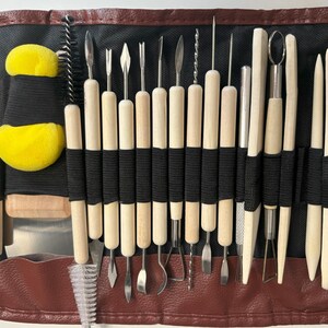 May include: A set of 28 pottery tools in a brown leather case. The tools are made of wood and metal and are used for shaping and sculpting clay. The case has a zipper closure and includes a small brush and two yellow sponges.