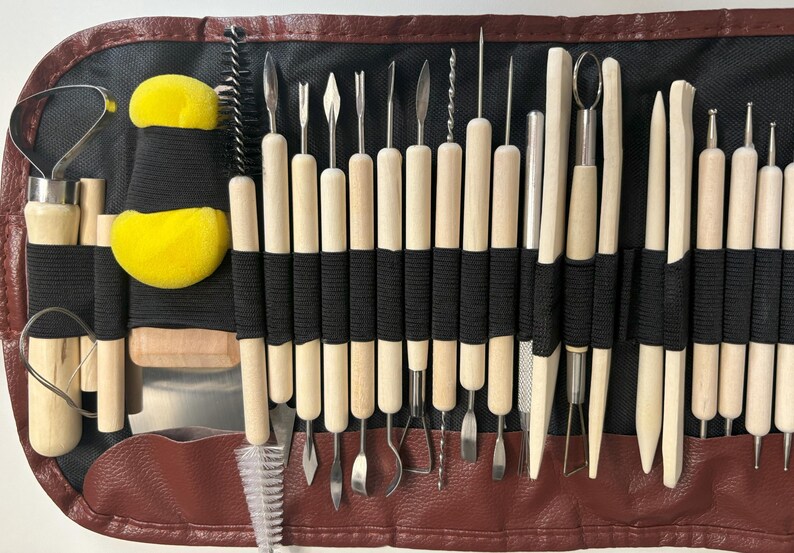 May include: A set of 28 pottery tools in a brown leather case. The tools are made of wood and metal and are used for shaping and sculpting clay. The case has a zipper closure and includes a small brush and two yellow sponges.