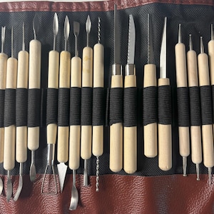 Pode incluir: Um conjunto de 20 ferramentas de escultura de argila com cabos de madeira e pontas de metal. As ferramentas est&atilde;o dispostas em um rolo de couro preto e marrom.