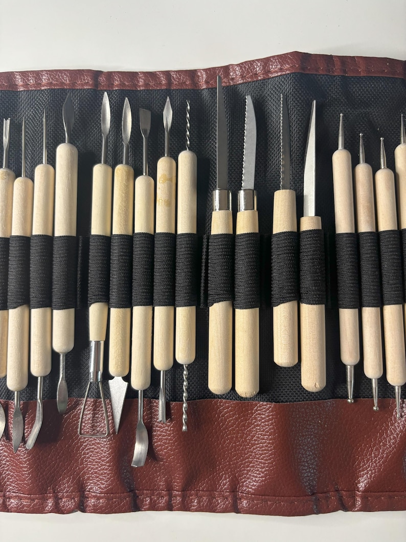 Pode incluir: Um conjunto de 20 ferramentas de escultura de argila com cabos de madeira e pontas de metal. As ferramentas est&atilde;o dispostas em um rolo de couro preto e marrom.