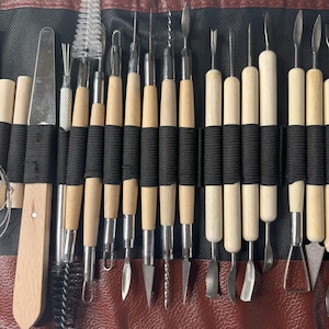 Pode incluir: Um conjunto de 20 ferramentas de escultura de argila com al&ccedil;as de madeira e v&aacute;rias pontas de metal. As ferramentas s&atilde;o enroladas em uma caixa de couro marrom com um z&iacute;per preto.
