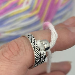 May include: A silver ring with a textured design is used to hold a white yarn tail. The yarn is pink and white.