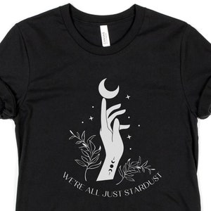 Może przedstawiać: Czarny T-shirt z białym nadrukiem przedstawiającym dłoń trzymającą p&oacute;łksiężyc z gwiazdami i liśćmi. Pod nadrukiem znajduje się tekst "We're all just stardust".