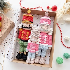 Può includere: Tre bambole schiaccianoci all'uncinetto in una scatola. Le bambole hanno uniformi rosse, rosa e grigie con baffi neri e capelli bianchi. La scatola è foderata con carta festiva con motivi di cervi e alberi.