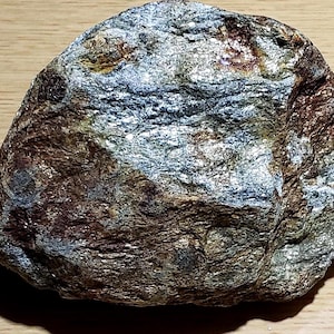 May include: A rough, irregularly shaped rock featuring a mix of metallic silver, deep brown, and earthy green tones. The surface displays a layered, crystalline texture with visible mineral inclusions and reflective patches across its uneven face.
