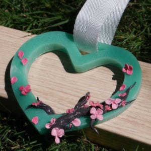 May include: A teal heart-shaped ornament with pink cherry blossoms and petals. The ornament has a white ribbon for hanging. The blossoms and petals are scattered across the heart, with a dark branch design.