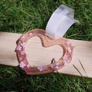 May include: A heart-shaped ornament made of translucent pink resin, adorned with small pink flowers and vines. The ornament has a glittery interior and a white ribbon for hanging. The background is a wooden surface and green grass.