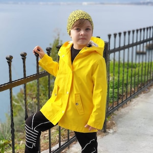 May include: A young person wearing a yellow raincoat with a hood, black trousers with white accents, and a yellow and black patterned hat. The person is standing next to a black metal fence.