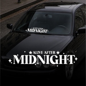 Op de afbeelding: Een zwarte auto met een witte sticker op de voorruit met de tekst "ALIVE AFTER MIDNIGHT" in een gestileerd lettertype. De tekst wordt ook op de motorkap herhaald. De auto staat geparkeerd op straat.