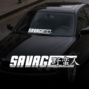 Könnte beinhalten: Weißer "SAVAGE" Autoaufkleber mit japanischen Schriftzeichen, auf der Windschutzscheibe eines schwarzen Autos. Der Aufkleber hat eine fette, serifenlose Schrift. Das Auto steht auf einer dunklen Straße.