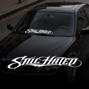 Op de afbeelding: Witte vinyl auto sticker met de tekst "Still Hated" in een gestileerd script. De sticker is geplaatst op de voorruit van een zwarte auto, met de motorkap en koplampen zichtbaar.