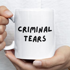 May include: A white ceramic mug with the words "CRIMINAL TEARS" in black, handwritten-style lettering. The mug is held by a person wearing a gold ring and bracelet.