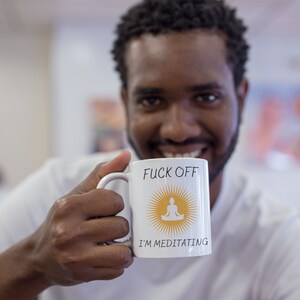 以下が含まれることがあります： 「F**k Off I'm Meditating」という文字と、白いシルエットの瞑想する人が描かれた黄色い太陽が描かれた白いセラミックマグカップ