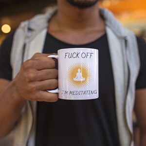 以下が含まれることがあります： 「F*ck Off I'm Meditating」という文字と、黄色の太陽の中に瞑想している人のグラフィックが描かれた白いセラミック製のコーヒーカップです。