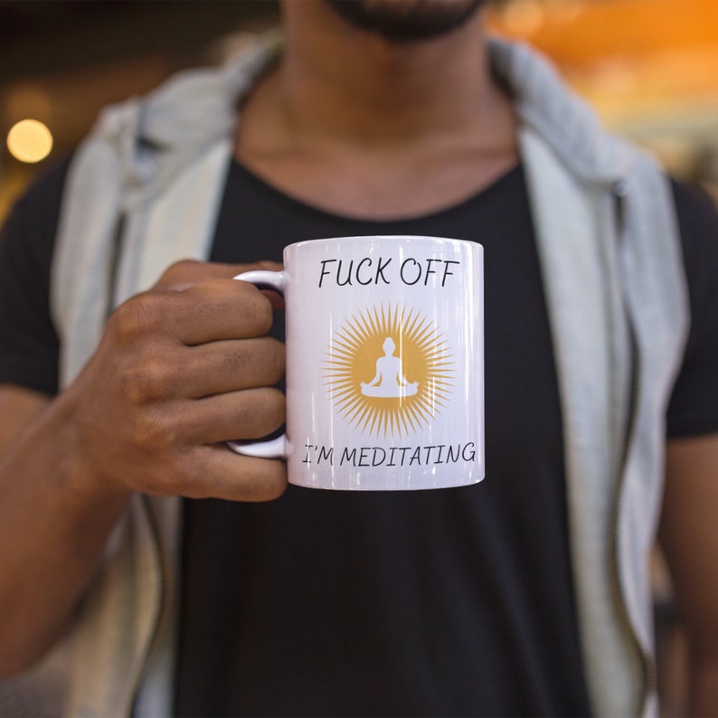 以下が含まれることがあります： 「F*ck Off I'm Meditating」という文字と、黄色の太陽の中に瞑想している人のグラフィックが描かれた白いセラミック製のコーヒーカップです。