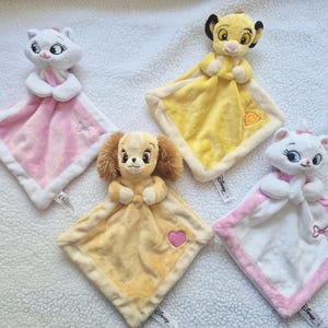 Peut inclure: Quatre doudous plats Disney composés de têtes de personnages attachées à des carrés de tissu doux. L'ensemble comprend deux doudous Marie le chat blancs avec des accents roses, un doudou Simba le lion jaune et un doudou Lady le chien beige avec un cœur rose.