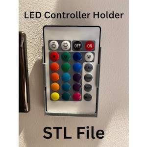 May include: A 3D printed holder for an LED controller with buttons for on/off, flash, strobe, fade, and smooth. The controller has a row of colored buttons for red, green, blue, and white. The holder is gray and has the text "STL File" printed below it.