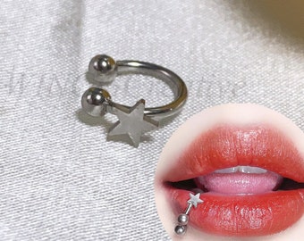 May include: A silver-colored horseshoe lip ring with a star charm and two ball ends. A small image shows a person wearing a lip piercing with dangling star charms. The background is a white, textured fabric.