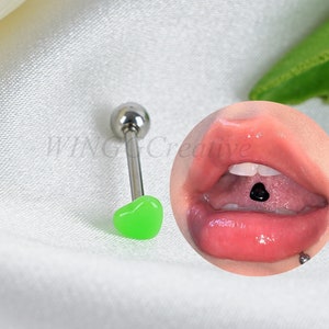 Puede incluir: Una barra de piercing en forma de corazón verde con una bola plateada en un extremo. La barra se muestra sobre una superficie blanca con un primer plano de una lengua con un piercing de barra negra.
