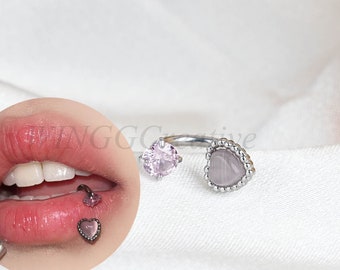 May include: A silver ring with a heart-shaped stone and a smaller heart-shaped stone on the side. The ring is on a white surface.