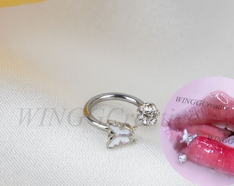 May include: Silver butterfly nose ring with a clear crystal ball on the end. The ring is shown on a white background with a close-up of a pair of lips wearing the ring.