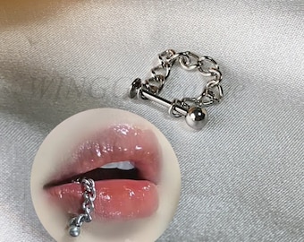 May include: Silver chain lip ring with a small ball at the end. The chain is attached to a bar that goes through the lip. The ring is shown on a model's lip.