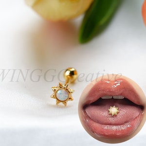 May include: Gold tongue ring with a white opal stone in the center and a sun design. The ring is shown on a person's tongue.