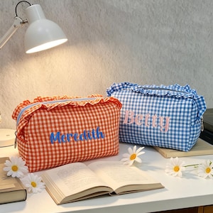 Puede incluir: Dos estuches rectangulares con cremallera, bordes con volantes y estampado de cuadros vichy. Uno es naranja y blanco con el nombre Meredith bordado en azul, y el otro es azul y blanco con el nombre Betty bordado en rosa claro.