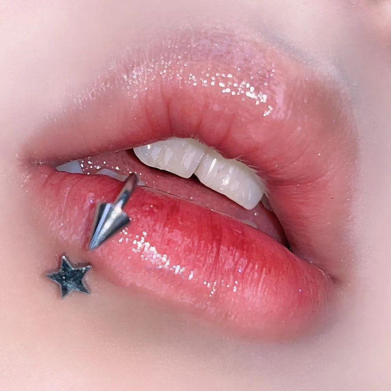 Peut inclure: Gros plan sur les l&egrave;vres d'une personne avec un piercing labial en forme d'&eacute;toile et de pointe argent&eacute;e. Les l&egrave;vres sont brillantes et roses.