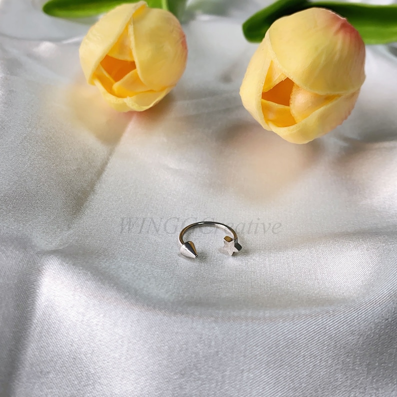 Peut inclure: Une bague en argent avec une &eacute;toile et une pointe pointue &agrave; chaque extr&eacute;mit&eacute;. La bague est sur un fond de tissu blanc avec deux tulipes jaunes.
