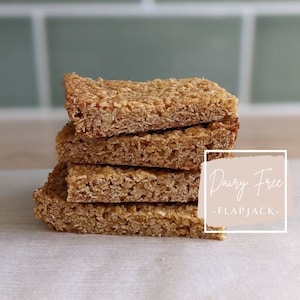 Può includere: Una pila di quattro flapjacks senza latticini, a base di avena e miele, su una superficie bianca. Il testo "Dairy Free - Flapjack -" è scritto su un rettangolo rosa.