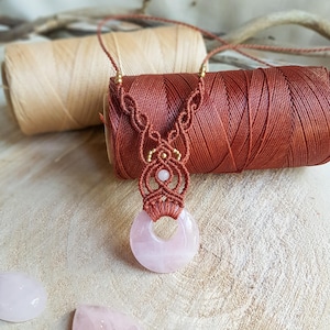 Può includere: Una collana con un ciondolo in quarzo rosa, un design macramé e perline dorate. La collana è realizzata con un cordoncino marrone ed è esposta su un rocchetto di filo marrone.