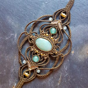 Puede incluir: Un collar de macramé marrón con una gran piedra preciosa ovalada verde y piedras preciosas redondas verdes más pequeñas. El collar está decorado con cuentas doradas.