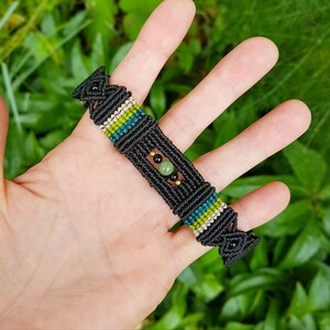 Peut inclure: Bracelet macramé noir avec des accents verts, turquoise et blancs. Le bracelet présente un motif central avec une pierre verte et des perles noires. Le macramé est noué selon un motif géométrique, créant un aspect texturé. Un accessoire fait main unique.