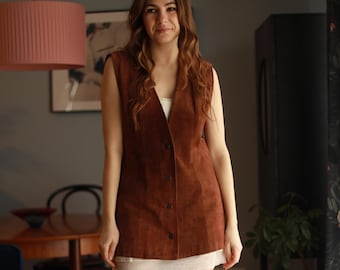 May include: A woman wearing a brown suede vest over a white top and a short skirt. She is also wearing brown boots. The vest has a button-up front. The outfit is in a room with a blue sofa and a wooden table.